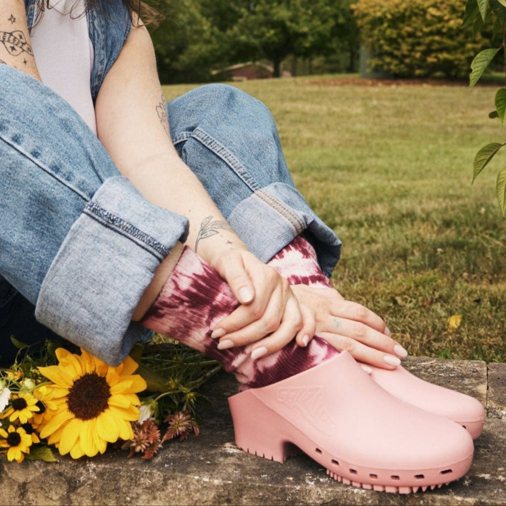 Person wearing pink clogs with a bouquet of flowers and denim jeans in a garden setting