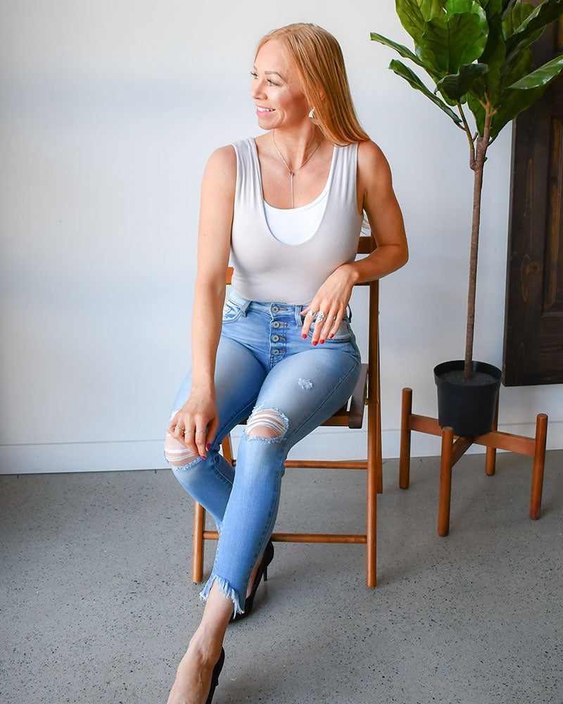 Woman sitting on a chair wearing a white tank top and light blue jeans with a plant in the background.