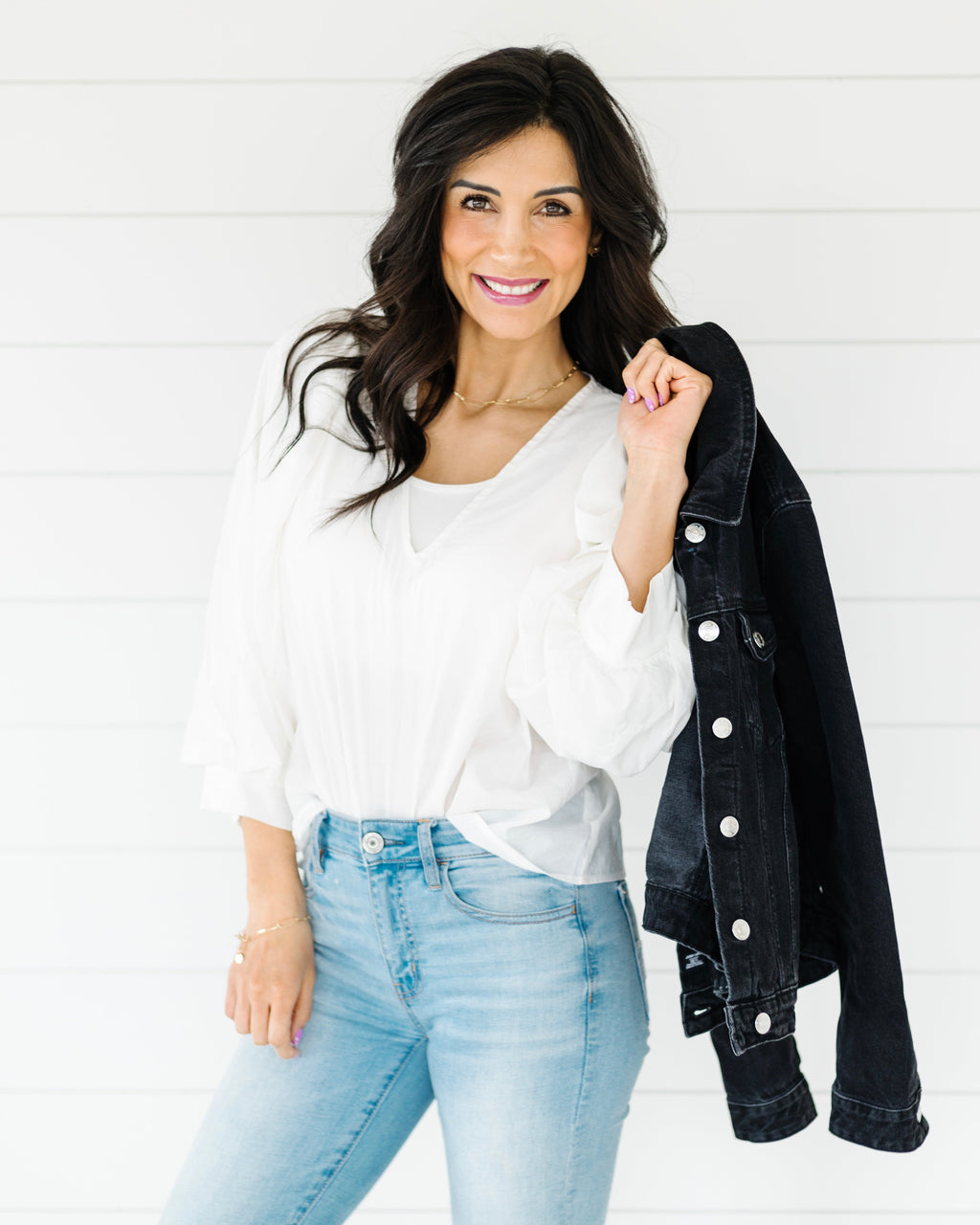 Woman wearing a white blouse, light blue jeans, and a black denim jacket against a white background