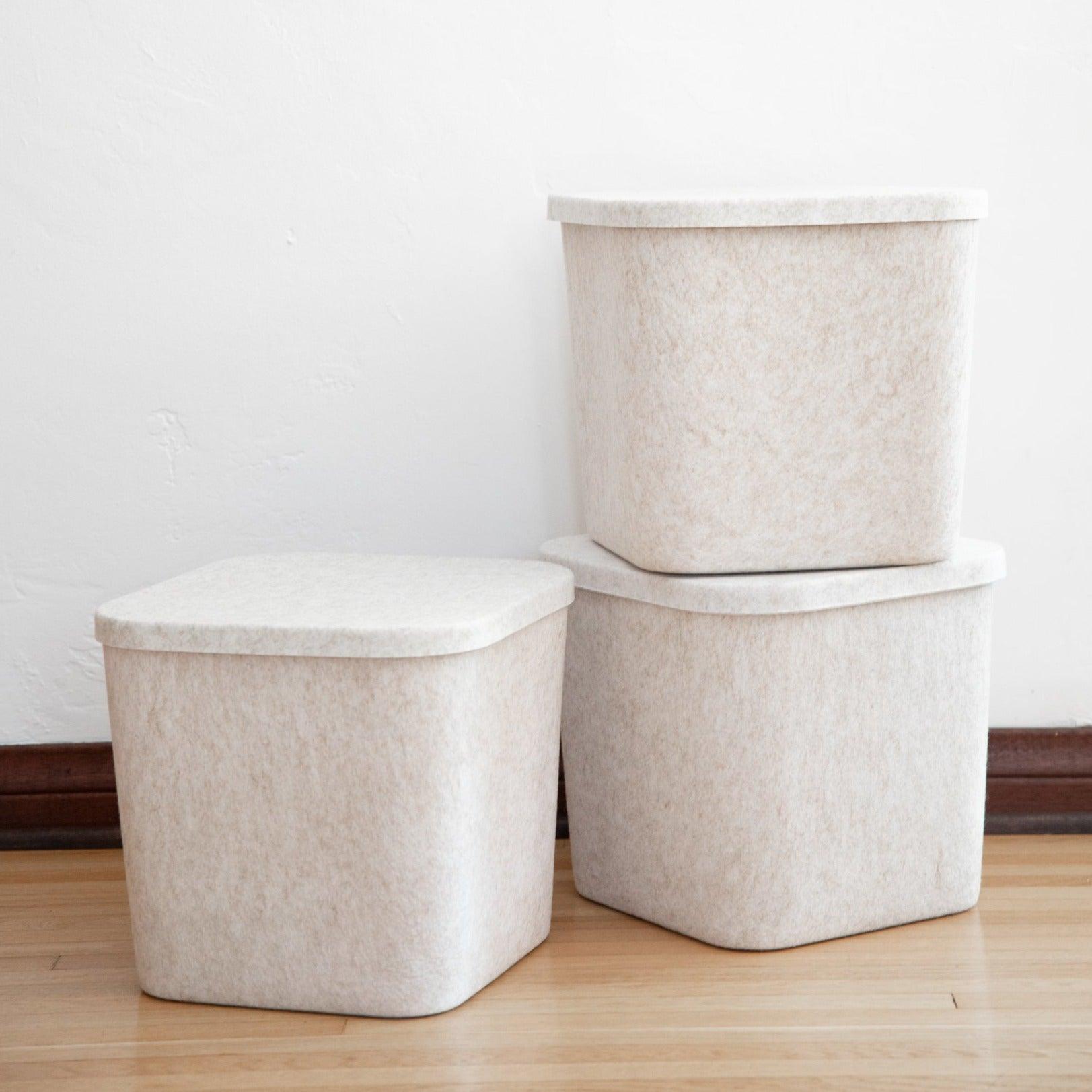 Three beige storage bins with lids on a wooden floor against a white wall.