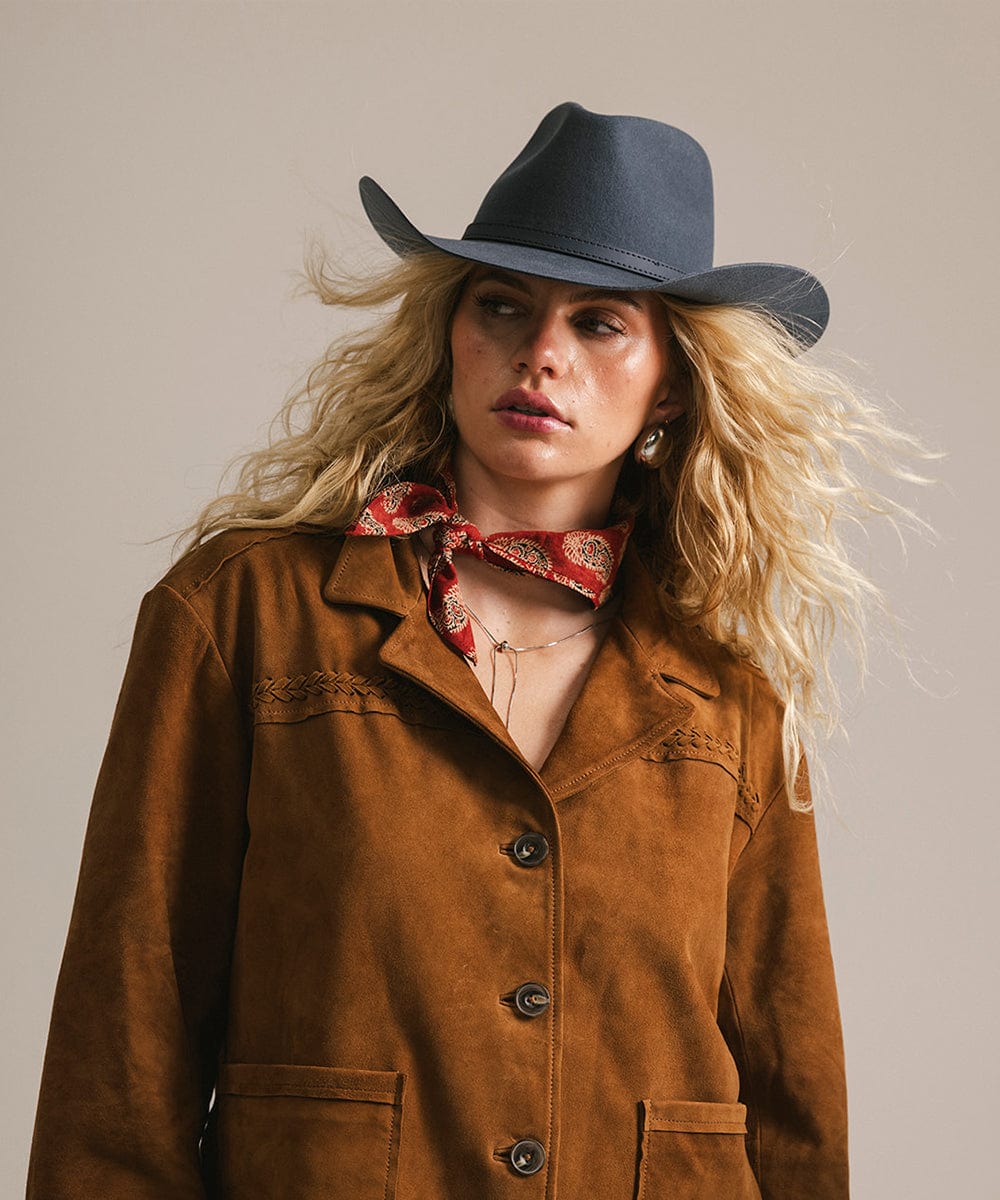 Woman wearing a brown jacket, black cowboy hat, and red bandana against a neutral background
