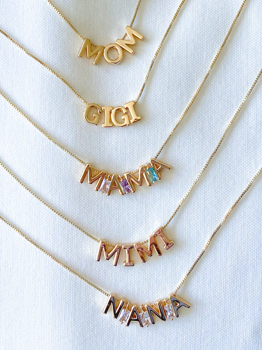 Gold necklaces with 'MOM', 'GIGI', 'MAMA', 'MIMI', and 'NANNA' charms on a white background.