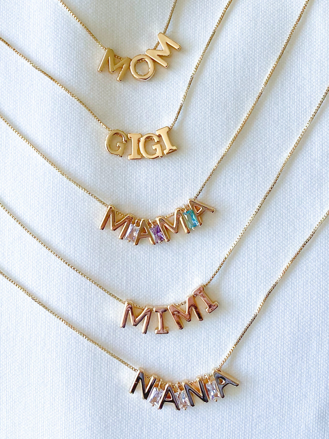 Gold necklaces with 'MOM', 'GIGI', 'MAMA', 'MIMI', and 'NANNA' charms on a white background.