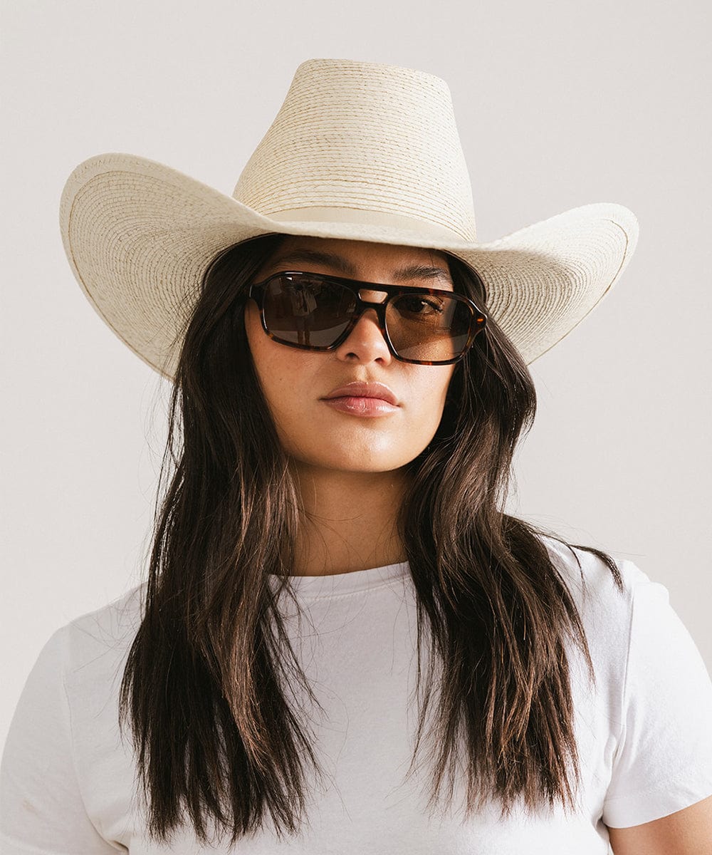 Woman wearing a straw cowboy hat and sunglasses on a plain background