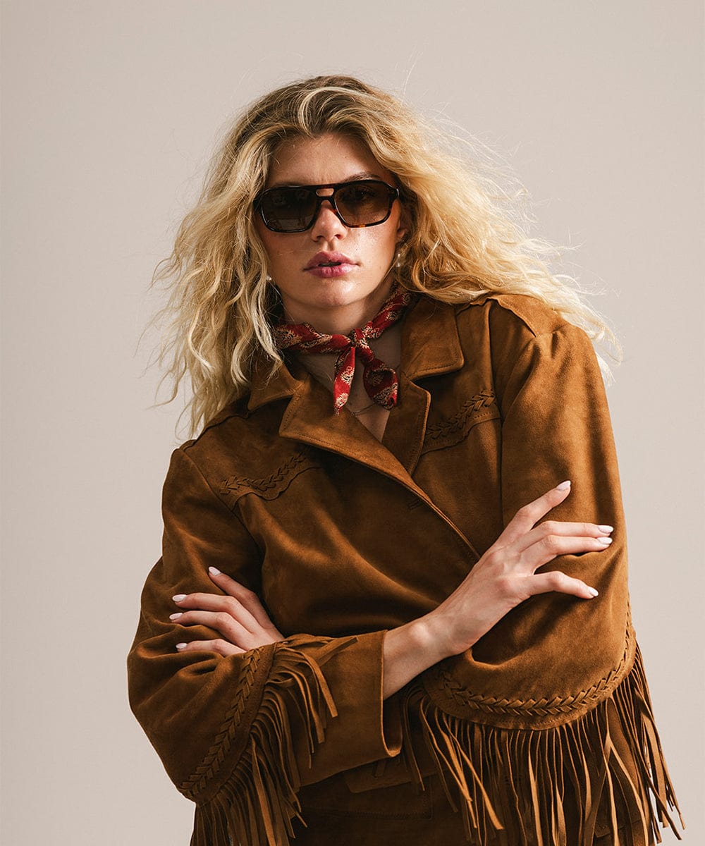 Woman wearing a brown fringed jacket and sunglasses against a plain background