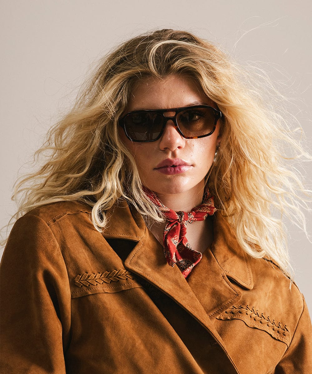Woman wearing sunglasses and a brown jacket with a red scarf, against a neutral background