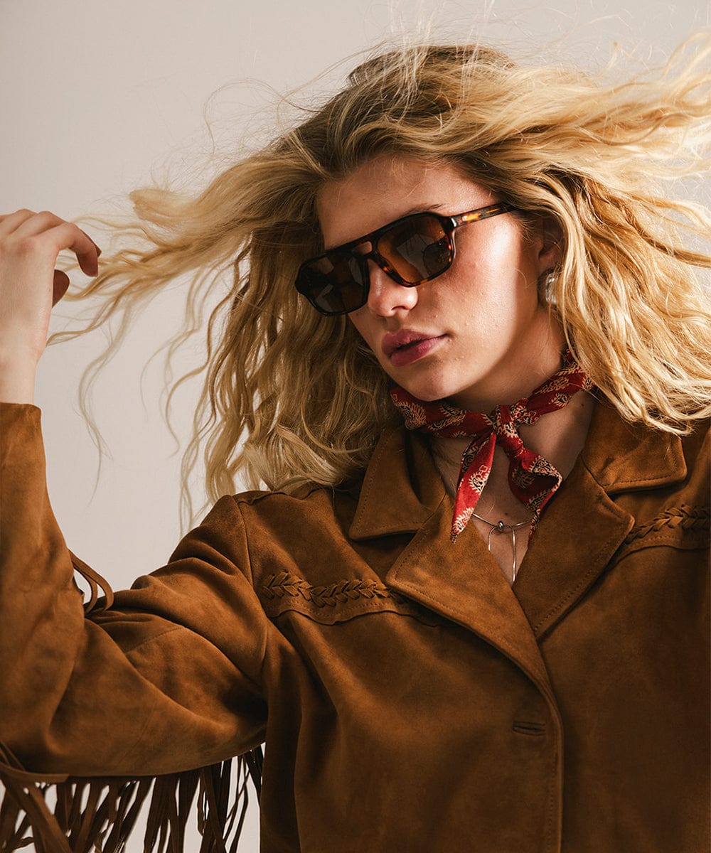 Woman wearing sunglasses and a brown coat with a red scarf, against a neutral background