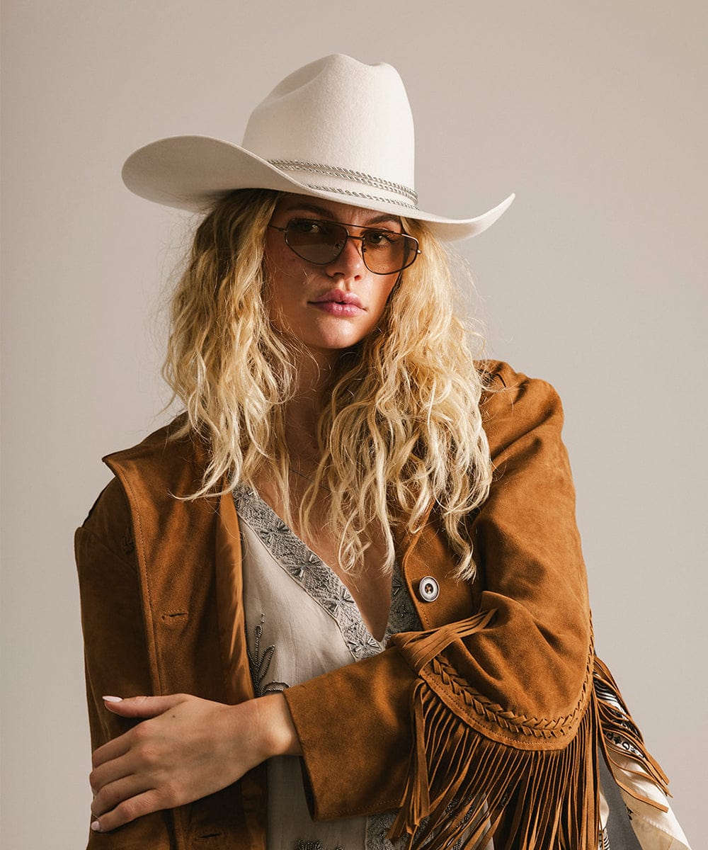 Woman wearing a brown suede jacket with fringe, white cowboy hat, and sunglasses on a plain background