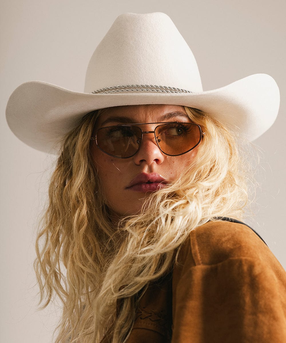 Person wearing a white cowboy hat and sunglasses with a neutral background