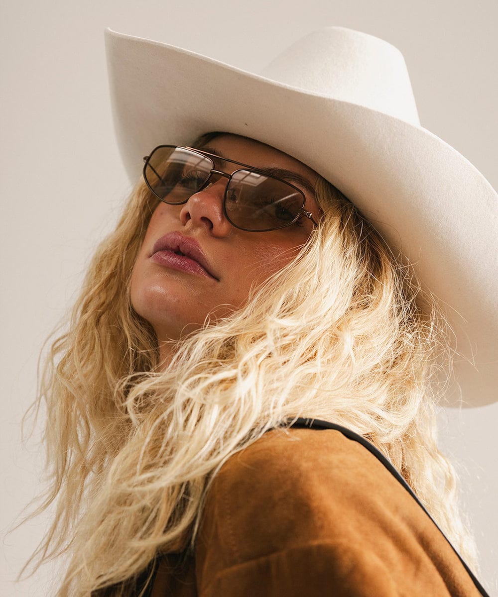 Person wearing a white cowboy hat and sunglasses with a neutral background