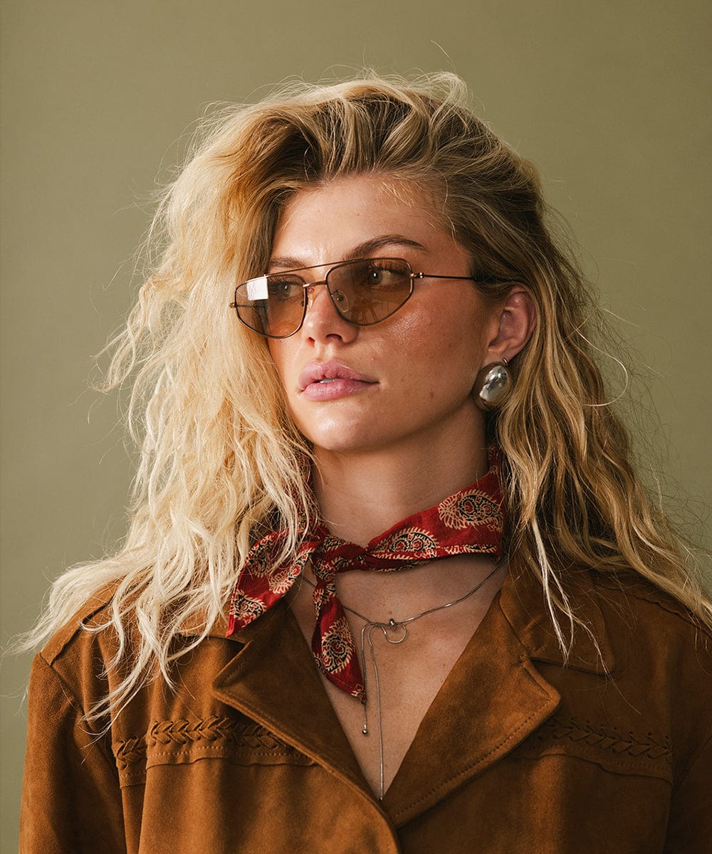 Woman wearing sunglasses and a brown jacket with a red scarf against a green background
