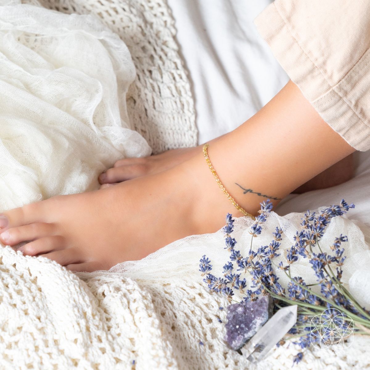 Close-up of a foot with a gold anklet on a white textured surface with lavender flowers.