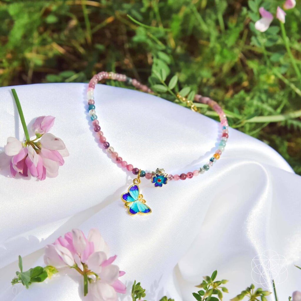 Colorful beaded necklace with a butterfly charm on a white surface with pink flowers and greenery.