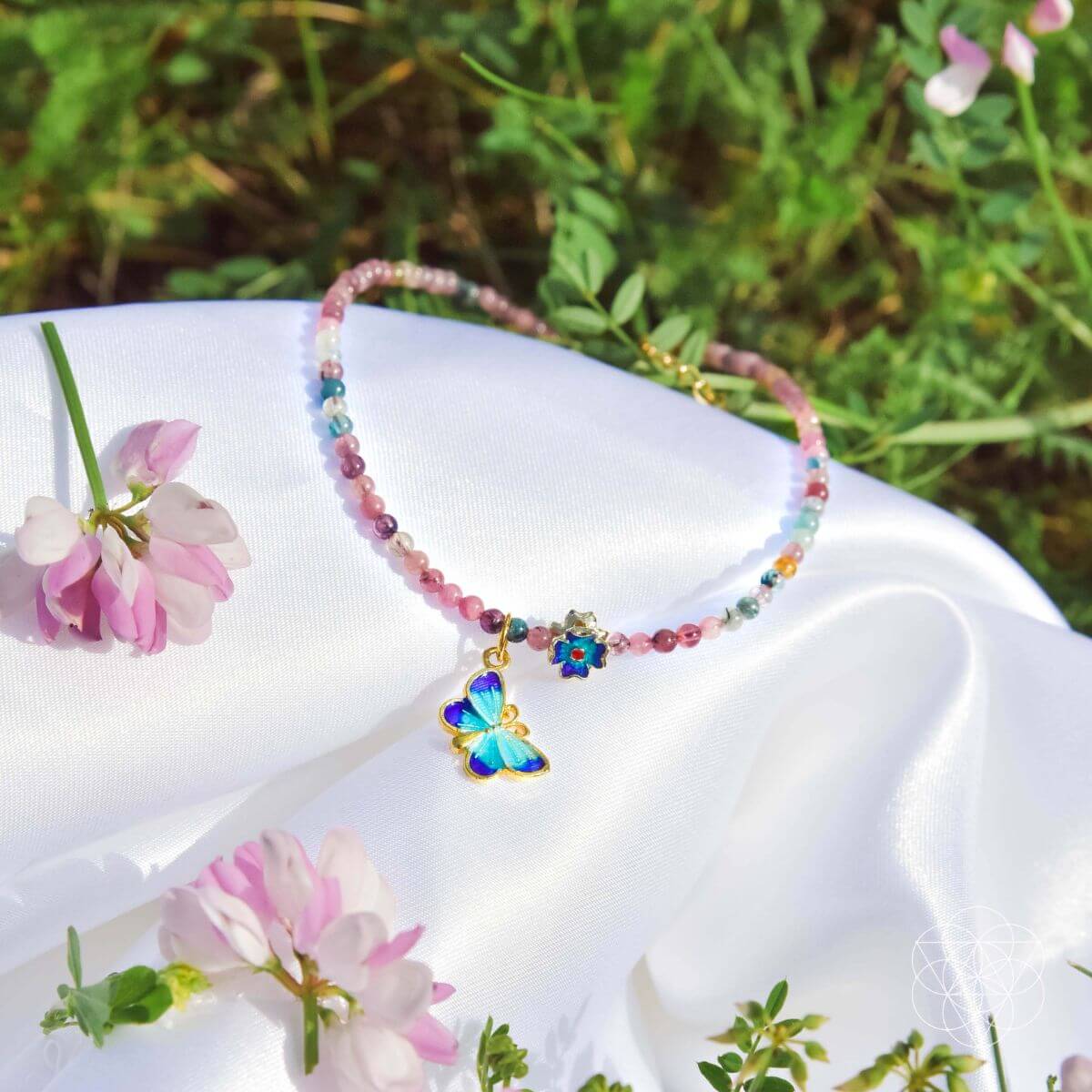 Colorful beaded necklace with a butterfly charm on a white surface with pink flowers and greenery.