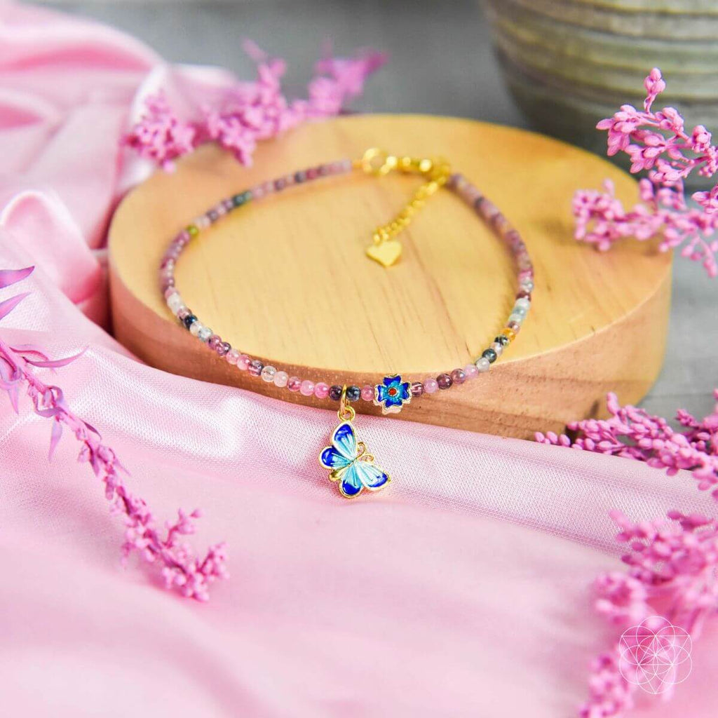 Colorful beaded bracelet with a butterfly charm on a wooden stand, surrounded by pink flowers.