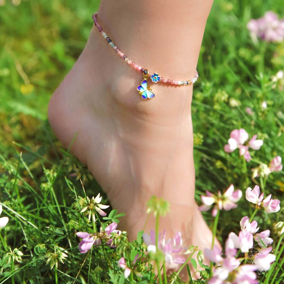 Ankle bracelet with a colorful flower charm worn on a foot in a grassy field with flowers.